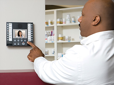 From the secured side of a pharmacy door, a pharmacist is using an exchange-based video intercom master mounted to a wall to clearly see and speak with a nurse or doctor requesting entry