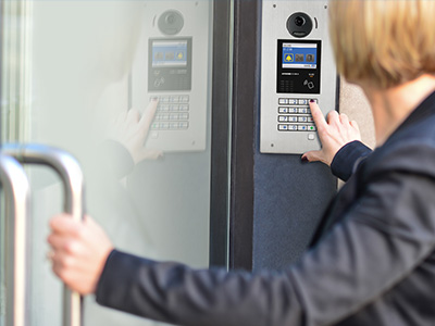 Self contained stainless steel multi-tent entry unit is flush mounted on the mullion of a glass apartment door while a female resident enters an access code to be allowed building entry.